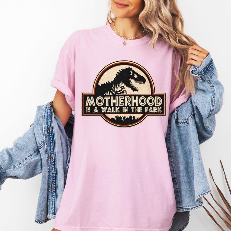 May include: A light pink t-shirt with the text "MOTHERHOOD IS A WALK IN THE PARK" in a black and brown graphic. The graphic features a dinosaur silhouette inside a circular frame.