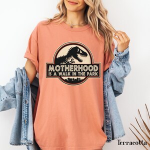 May include: Terracotta-colored t-shirt featuring a dinosaur graphic within a circular emblem. The emblem reads "Motherhood is a Walk in the Park" in a bold, stylized font. The shirt has short sleeves and a relaxed fit.
