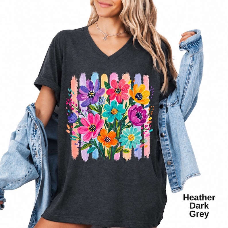 Op de afbeelding: Een donkergrijs gem&ecirc;leerd T-shirt met V-hals en een kleurrijke bloemenprint in paars, roze, geel en turquoise, geplaatst tegen een achtergrond van verticale pastelkleurige penseelstreken. Het shirt wordt gedragen met een lichtblauw spijkerjasje.