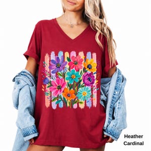 Op de afbeelding: Een heather kardinaalrood T-shirt met V-hals voorzien van een levendige grafische print van kleurrijke geschilderde bloemen in paars, roze, geel en turquoise, geplaatst over verticale penseelstreken in pasteltinten. Een lichtblauw spijkerjack.