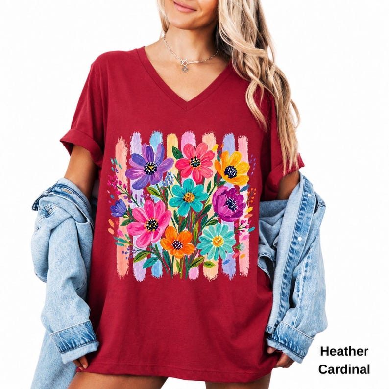 Op de afbeelding: Een heather kardinaalrood T-shirt met V-hals voorzien van een levendige grafische print van kleurrijke geschilderde bloemen in paars, roze, geel en turquoise, geplaatst over verticale penseelstreken in pasteltinten. Een lichtblauw spijkerjack.