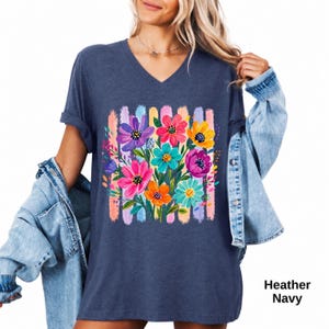 Op de afbeelding: Een heather navy V-hals t-shirt met een levendige grafische print van kleurrijke bloemen in tinten roze, geel, oranje, paars en taling. Het bloemmotief is geplaatst over verticale penseelstreekstrepen in zachte pastelkleuren.