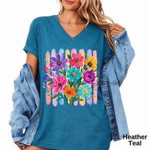 Op de afbeelding: Een heather teal V-hals t-shirt met een levendige grafische print van een kleurrijk bloemenboeket. De bloemen zijn geschilderd in tinten roze, paars, geel en lichtblauw, geplaatst tegen een achtergrond van verticale pastelkleurige penseelstreken.