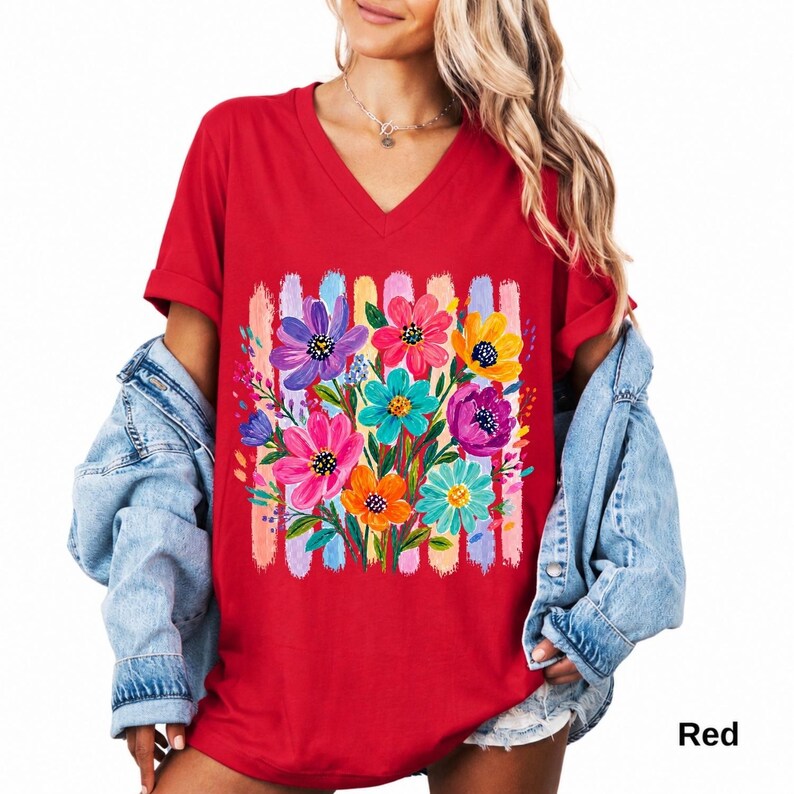 Op de afbeelding: Een rood T-shirt met V-hals voorzien van een centrale afbeelding van een kleurrijk boeket geschilderde bloemen in roze, paars, geel en turquoise, tegen een achtergrond van verticale penseelstreken in pastelkleuren.