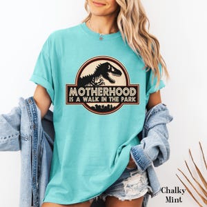 May include: A Chalky Mint t-shirt with a graphic that reads "MOTHERHOOD is a WALK IN THE PARK". The graphic features a dinosaur silhouette inside a circular emblem with a brown border. The shirt has rolled-up sleeves.