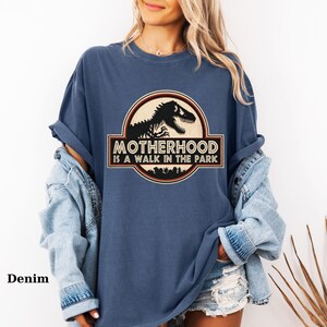 May include: Denim blue t-shirt with a graphic that reads "Motherhood is a Walk in the Park." The graphic features a black dinosaur silhouette inside a circular emblem with a brown border. The shirt has rolled-up sleeves.