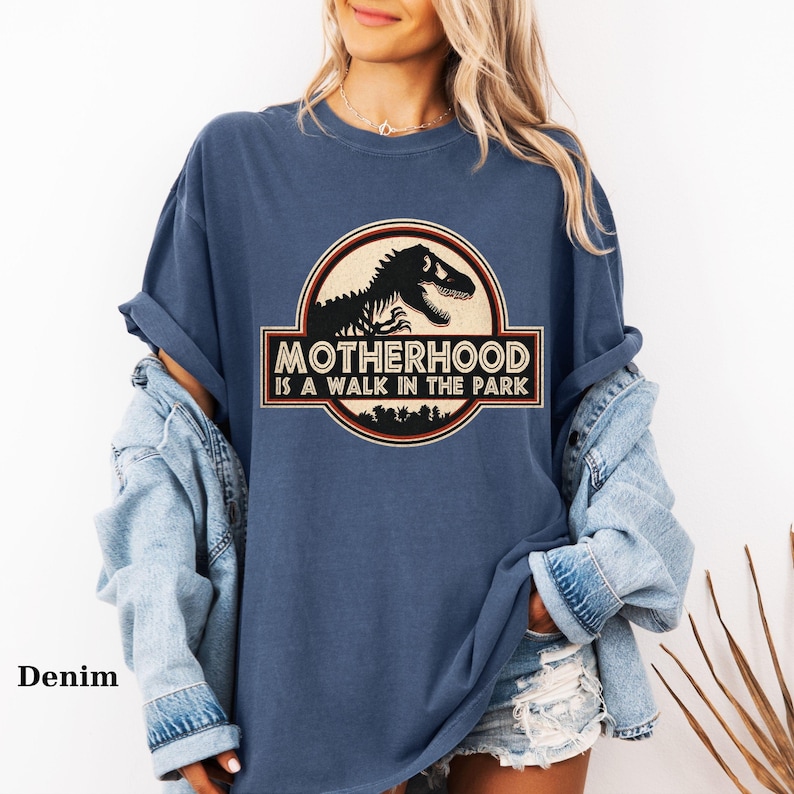 May include: Denim blue t-shirt with a graphic that reads "Motherhood is a Walk in the Park." The graphic features a black dinosaur silhouette inside a circular emblem with a brown border. The shirt has rolled-up sleeves.