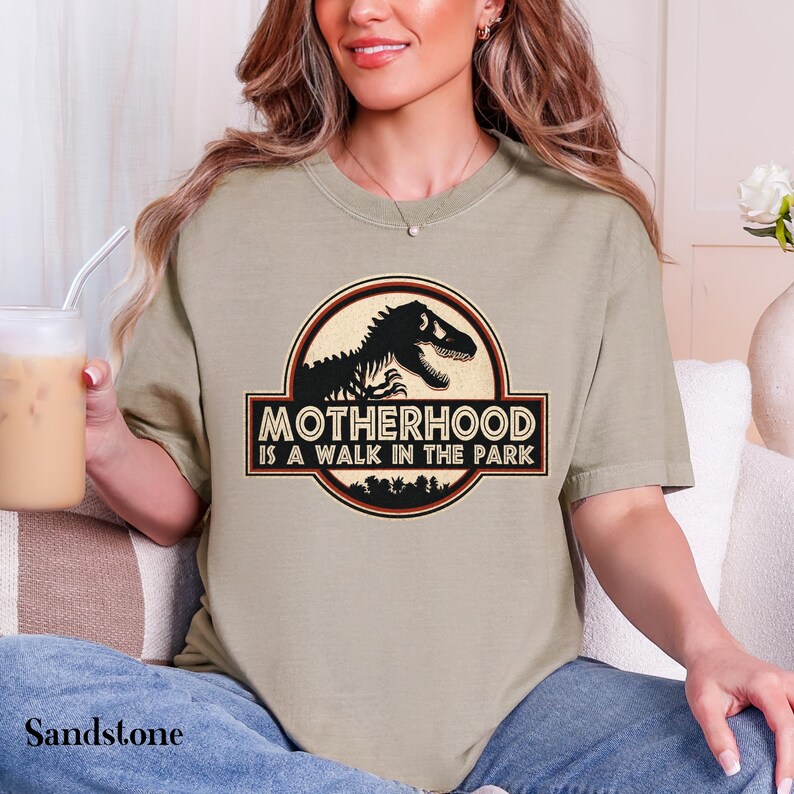 May include: Sandstone-colored t-shirt with a circular graphic featuring a black dinosaur silhouette and the text "MOTHERHOOD IS A WALK IN THE PARK" in a red and black design. The shirt is a short-sleeved crew neck.