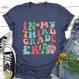 May include: Navy blue t-shirt with the text "IN MY THIRD GRADE ERA" in colorful, retro-style lettering. The design includes a book, daisy, pencil, and a colorful apple. Black and white sneakers are visible.