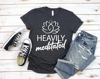 May include: A dark gray t-shirt with a white lotus flower graphic and the text "HEAVILY MEDITATED" in white. The shirt is laid out on a wooden surface with a pair of black canvas shoes and a pair of blue jeans.