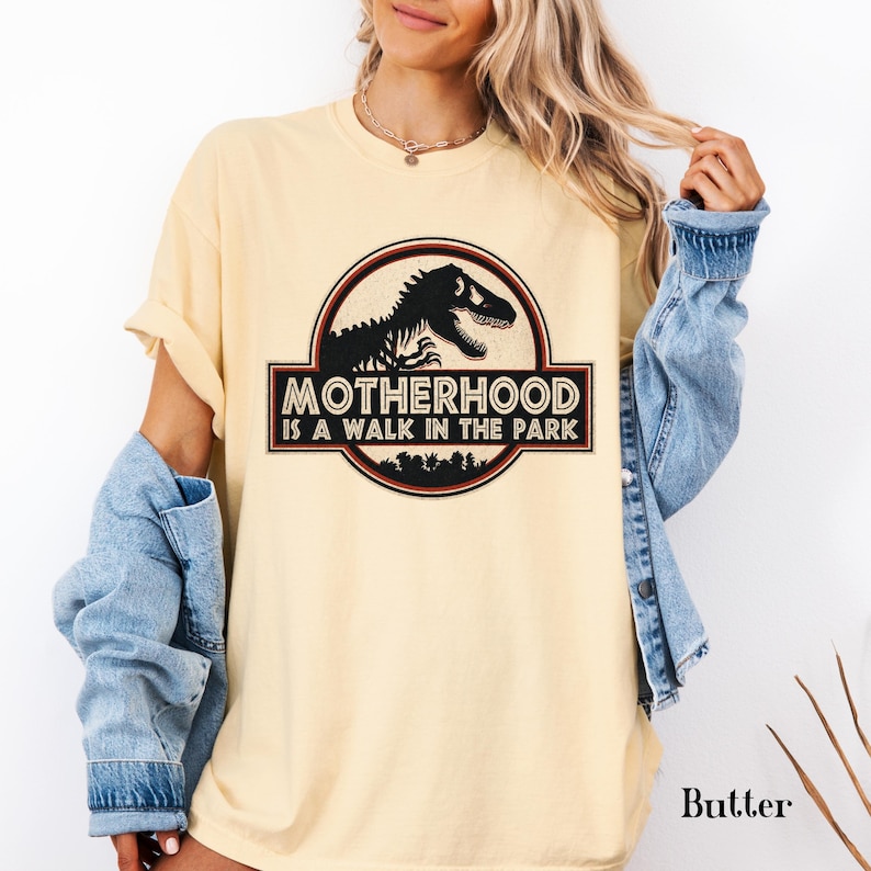 May include: Butter-colored t-shirt with a graphic that reads "Motherhood is a Walk in the Park." The design features a dinosaur silhouette inside a circular emblem with a red and black border. The shirt has short sleeves and a relaxed fit.