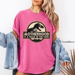 May include: A pink t-shirt with a graphic that reads "Motherhood is a Walk in the Park" with a dinosaur silhouette. The shirt has rolled-up sleeves and is paired with a denim jacket.