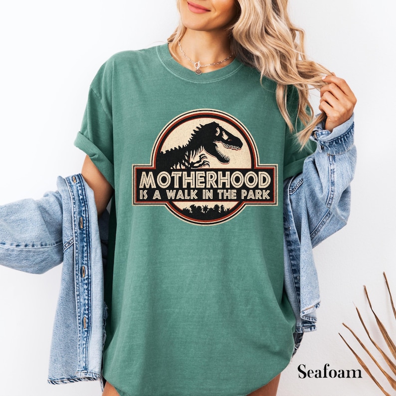 May include: Seafoam green t-shirt with a graphic featuring a dinosaur silhouette inside a circular emblem. The text "MOTHERHOOD IS A WALK IN THE PARK" is displayed below the dinosaur image. The shirt has rolled-up sleeves.