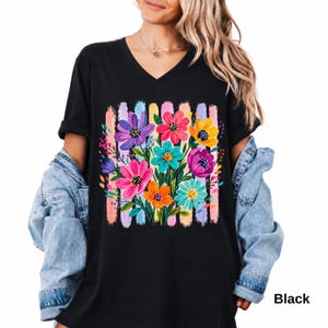 Op de afbeelding: Een zwart T-shirt met V-hals voorzien van een levendige grafische print van een kleurrijk bloemenboeket. De bloemen zijn uitgevoerd in tinten paars, roze, oranje, geel en turquoise, tegen een achtergrond van verticale, getextureerde pastelstreken.