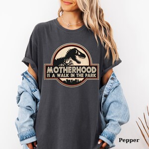 May include: Dark grey t-shirt with a circular graphic featuring a dinosaur silhouette and the text "MOTHERHOOD IS A WALK IN THE PARK". The shirt has rolled-up sleeves and is worn with a denim jacket. The word "Pepper" is printed at the bottom right.