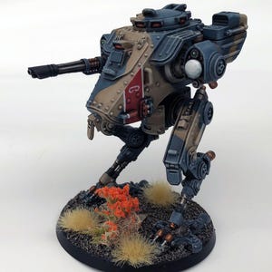 May include: A miniature, detailed model of a bipedal war machine, painted in shades of blue, tan, and red. The model features a cannon, intricate mechanical details, and is mounted on a circular base with miniature foliage.