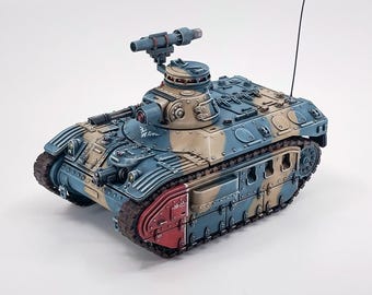 May include: A miniature tank model, painted in blue, tan, and red camouflage. The model has a turret with a gun, tracks, and detailed features. The tank is set against a white background.
