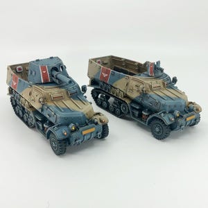 May include: Two miniature military vehicles, one with a cannon and the other an open-top transport. Both are painted in a camouflage pattern of tan, blue, and red accents. The vehicles have detailed tracks and wheels.