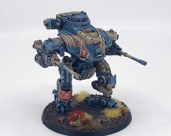 May include: A miniature, painted war machine model. The model is primarily blue and tan with red and white accents. It has two cannons and is mounted on a circular base with faux grass and foliage. The number 7 is visible on the side.
