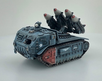 May include: A detailed model of a blue-grey military vehicle with four missiles. The vehicle has a red and grey base, a rotating turret, and intricate detailing. The missiles have grey bodies and red tips.