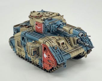 May include: A miniature model tank in a desert camouflage color scheme. The tank features a tan, blue, and red color palette with intricate detailing. The model has a large turret and multiple gun barrels, showcasing a military theme.