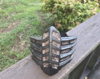May include: A black, intricately designed mask with a textured surface. The mask features a series of horizontal bars and a central, vertical panel with a detailed pattern. The mask is resting on a wooden surface, with greenery in the background.