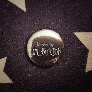 Könnte beinhalten: Ein glänzender, silberfarbener Knopf mit dem Text "Directed by Tim Burton" in einer gotischen Schriftart. Der Knopf befindet sich auf einem schwarzen Stoff mit einem weißen Sternmuster.