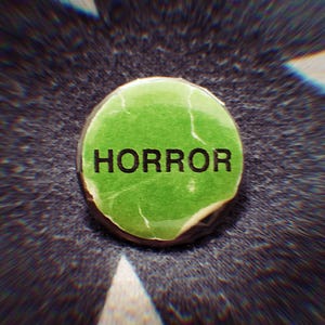 May include: A circular pinback button featuring a bright green background with the word HORROR printed in bold black capital letters across the center. The surface of the green paper insert shows visible creases and texture throughout the design.