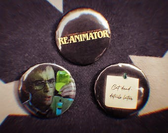 May include: Three round, black pinback buttons. One button has the text "RE-ANIMATOR" in gold. Another features a man in glasses holding a green liquid. The third has a note that reads "Cat dead, details later."