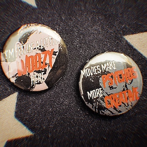 Könnte beinhalten: Zwei runde Ansteckbuttons mit Text. Einer sagt "I'M FEELIN A LITTLE WOOZY HERE" in Rot und Weiß auf schwarzem und rosa Hintergrund. Der andere sagt "MOVIES MAKE PSYCHOS MORE CREATIVE" in Rot und Weiß auf schwarzem Hintergrund.