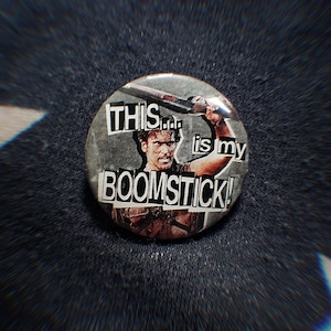 May include: A circular badge with the words "THIS... is my BOOMSTICK!" in a bold, cut-out style. The badge features a black and white image of a man holding a weapon. The background is a dark, textured fabric.