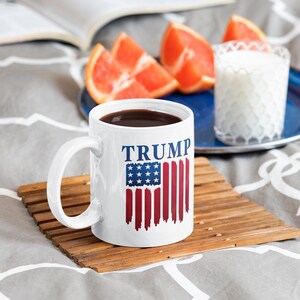 May include: White ceramic coffee mug with a blue and red American flag design and the word "TRUMP" printed in blue. The mug is sitting on a wooden coaster.