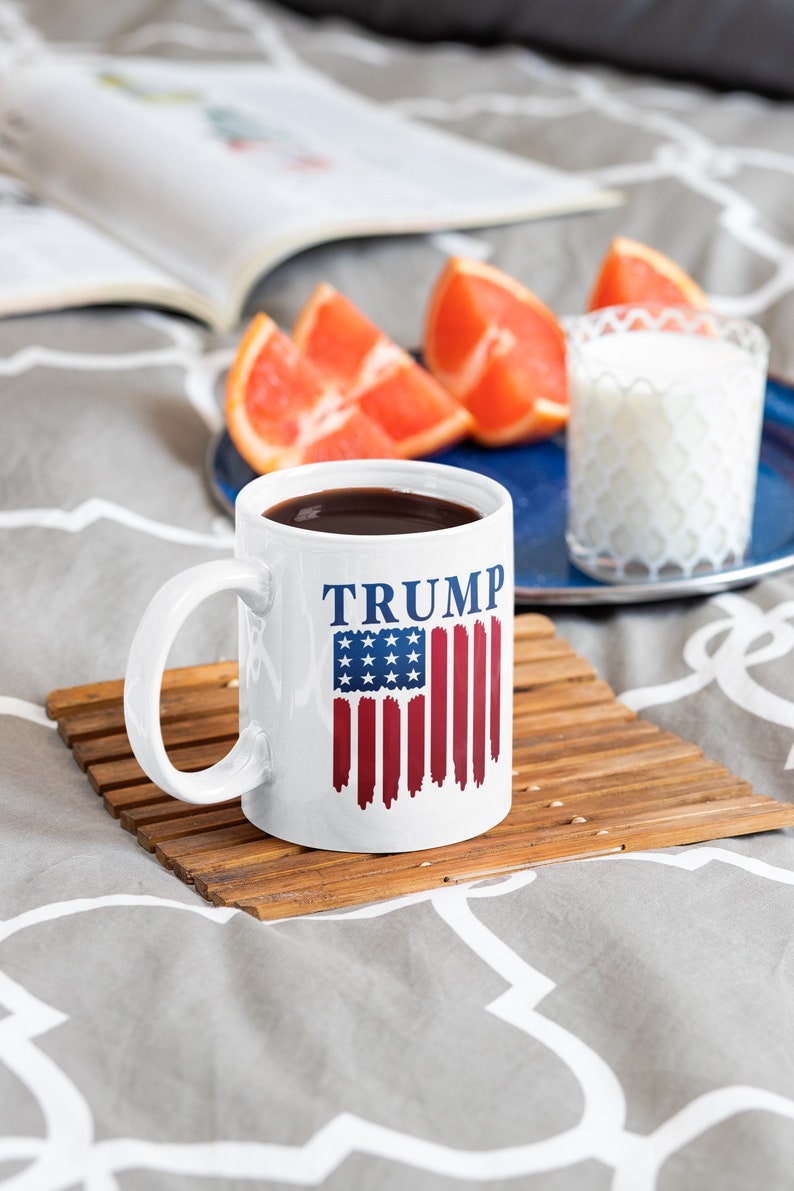 May include: White ceramic coffee mug with a blue and red American flag design and the word "TRUMP" printed in blue. The mug is sitting on a wooden coaster.