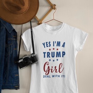 May include: White t-shirt with the text "Yes I'm a Trump Girl Deal with it!" in blue and red lettering with stars.