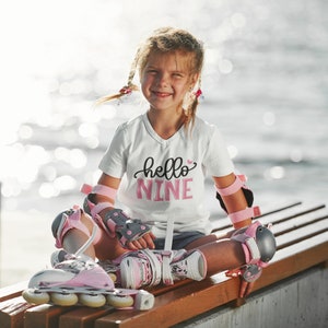 Puede incluir: Una ni&ntilde;a peque&ntilde;a con una camiseta blanca con el texto "hello nine" en letras rosas. Lleva rodilleras y patines rosas y grises. Est&aacute; sentada en un banco de madera.