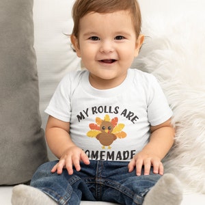 May include: A baby wearing a white onesie with a turkey graphic and the text "My rolls are homemade". The baby is sitting on a white fuzzy surface and wearing blue jeans.