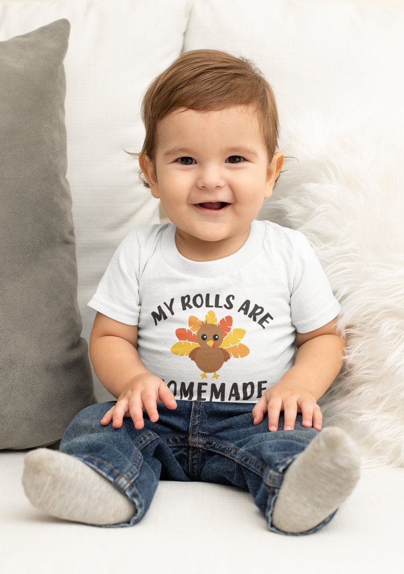 May include: A baby wearing a white onesie with a turkey graphic and the text "My rolls are homemade". The baby is sitting on a white fuzzy surface and wearing blue jeans.