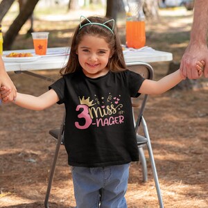 May include: A young girl wearing a black t-shirt with a pink number 3 and the text "Miss 3-Nager" in gold glitter. She is holding hands with two adults and smiling.