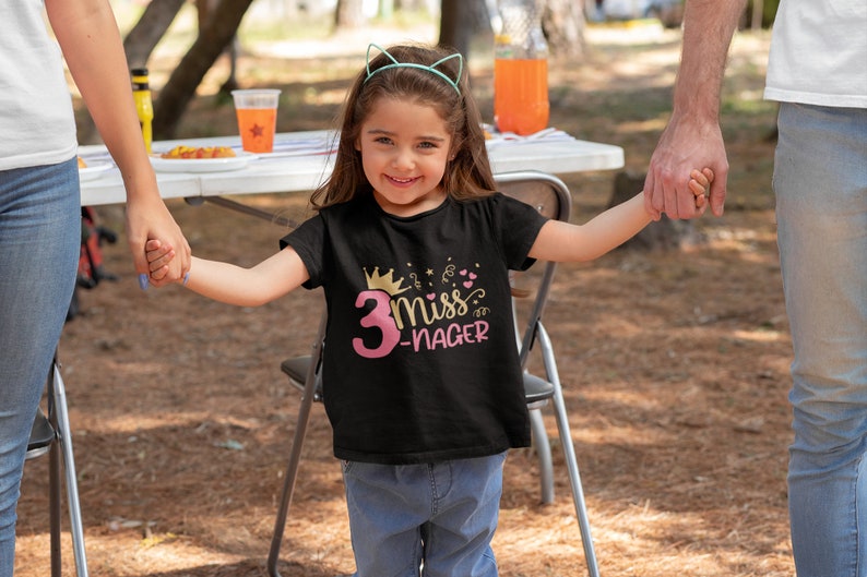 May include: A young girl wearing a black t-shirt with a pink number 3 and the text "Miss 3-Nager" in gold glitter. She is holding hands with two adults and smiling.