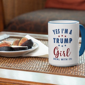 May include: White ceramic coffee mug with a blue rim and handle. The mug has red and blue text that reads "Yes I'm a Trump Girl Deal With It!" with stars between the words.