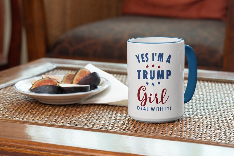 May include: White ceramic coffee mug with a blue rim and handle. The mug has red and blue text that reads "Yes I'm a Trump Girl Deal With It!" with stars between the words.