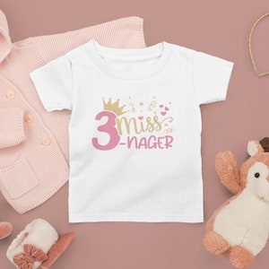 May include: White t-shirt with pink text that says "3 miss -nager" with a gold crown and pink hearts.