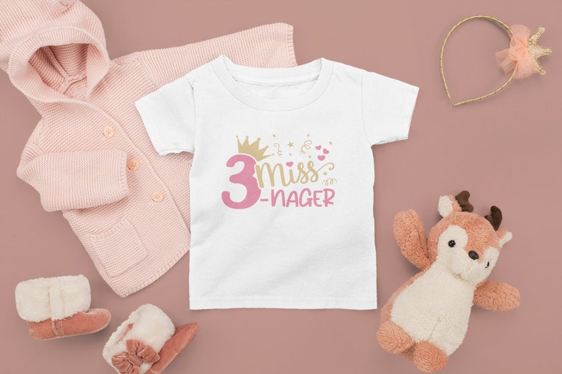 May include: White t-shirt with pink text that says "3 miss -nager" with a gold crown and pink hearts.