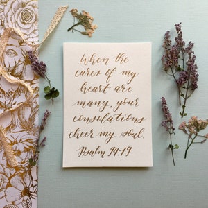 May include: A white card with gold calligraphy that reads "When the cares of my heart are many, your consolations cheer my soul. Psalm 94:19". The card is surrounded by dried flowers and lace on a light blue background.
