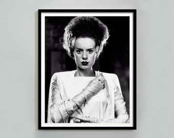 Peut inclure: Photographie en noir et blanc d'une femme avec une coiffure rappelant celle de Frankenstein. Elle porte une robe blanche et a une expression sérieuse.