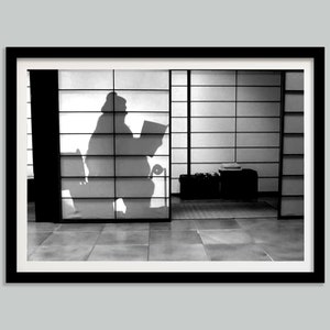 May include: Black and white photograph of a person's silhouette sitting in a room with a sliding door. The person is reading a book and their shadow is cast on the door.
