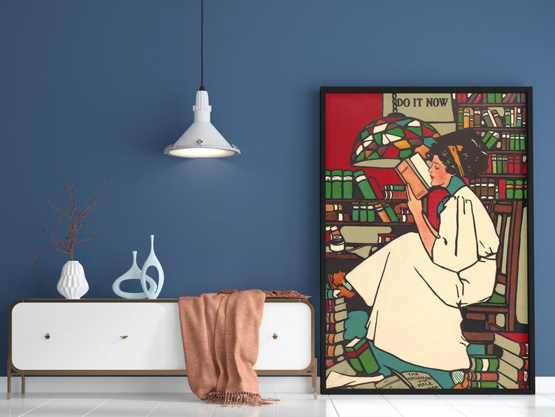 Puede incluir: Un p&oacute;ster enmarcado con una mujer leyendo un libro en un entorno de biblioteca. El p&oacute;ster tiene un fondo rojo con una pantalla de l&aacute;mpara de vidrio coloreado y el texto "DO IT NOW".