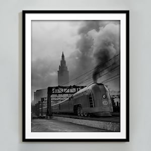 May include: A black and white photograph of a streamlined train pulling into a station. The train is emitting smoke and is in front of a tall building.