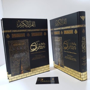 May include: Two black hardback books with gold accents and the title "The Holy Quran" on the front cover. The books feature a gold-coloured image of the Kaaba, a cube-shaped building in Mecca, Saudi Arabia, which is Islam's holiest place. The books also include the text "Colour Coded Tajweed Rules" and Arabic text. The books are sitting on a white surface.