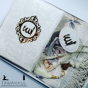 May include: A white prayer rug with a floral pattern, a white towel, a white prayer bead necklace, and a gold-coloured decorative piece with the word "Allah" in Arabic script.